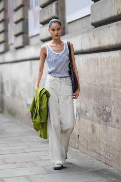 Model wears white and light blue striped tank top, white denim jean loose pants, bright green jacket in hand, dark brown leather bag, white boots...