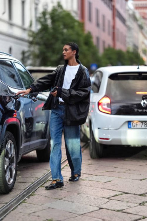 Model wears white cotton t-shirt, black oversized baggy leather jacket, shiny black woven leather bag, black leather belt, light blue denim jean...