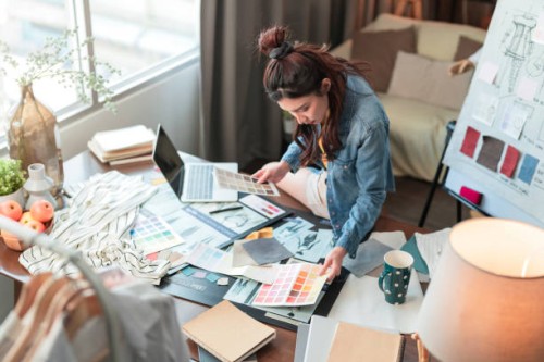 new work consulting on smartphone asian adult female woman fashion designer cloth tailor working for new collection sketch drawing choosing new color swatch fabric sample at her desk in her design studio home studio - fashion