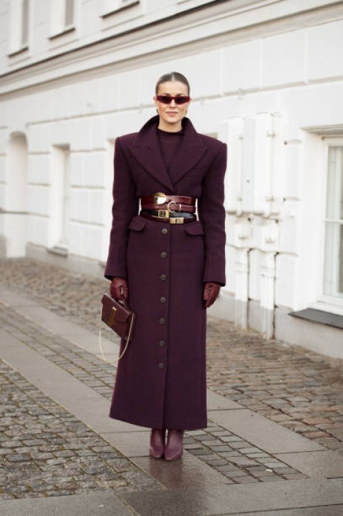 Nina Sandbech wears a burgundy top, long burgundy coat with multiple belts, burgundy bag, burgundy leather gloves and pointy burgundy shoes outside...