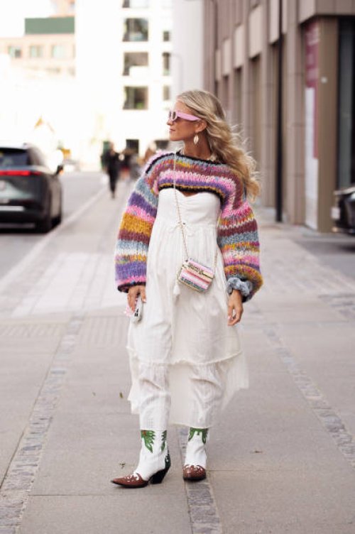 Olivia Anna-Catharina wears a long white dress, white green and brown cowboy boots, a colorful knitted cropped sweater, sunglasses with pink frame,...