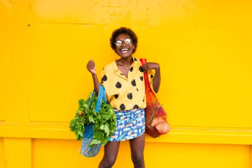 one young woman carrying grocery bags in front of yellow wall laughing - fashion stock pictures, royalty-free photos & images