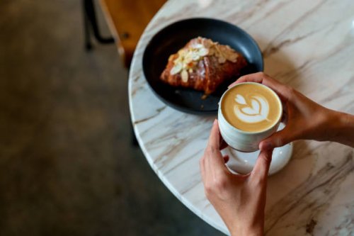 over the shoulder view of an asian woman enjoying her breakfast, a croissant with a cup of coffee in a cafe during the morning - coffee stockfoto's en -beelden