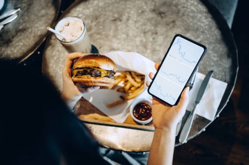 over the shoulder view of busy young woman checking financial trading data on smartphone while having her meal, beef and cheese burger with fries in a restaurant - junk food stock pictures, royalty-free photos & images