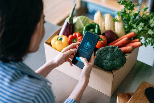 over the shoulder view of young asian woman doing home delivery grocery shopping online with mobile app device on smartphone at home, with a box of colourful and fresh organic vegetables and fruits on the table - food stock p