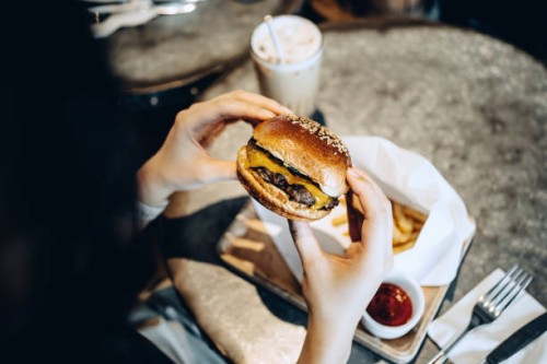 over the shoulder view of young woman enjoying her meal, eating a freshly made beef and cheese burger with fries and having a glass of iced milkshake in a restaurant - junk food stock pictures, royalty-free photos & images