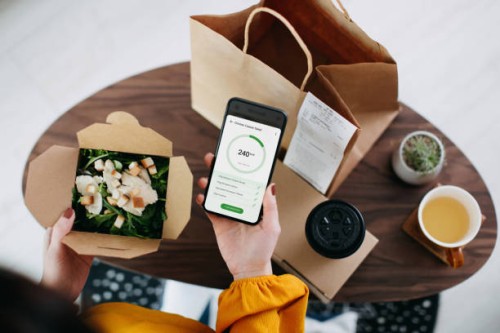 overhead view of young asian woman using health and fitness plan mobile app on smartphone to tailor make her daily diet meal plan, checking the nutrition facts and calories intake of the home delivery takeaway grilled chicken