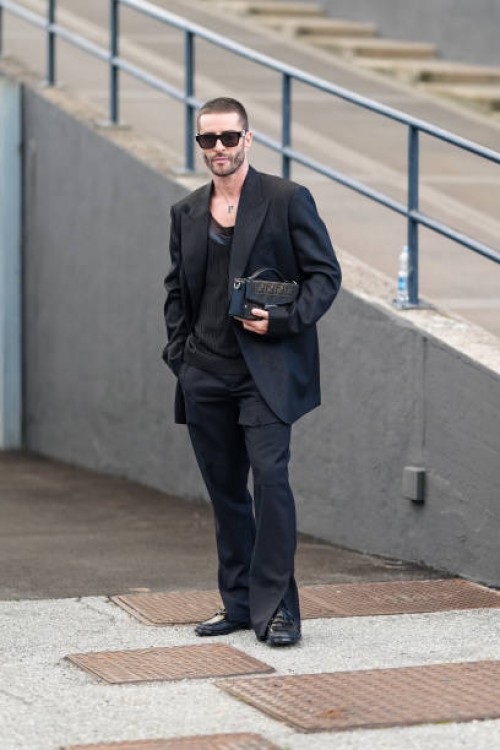 Pelayo Diaz wears black sunglasses, silver necklace, black oversized blazer jacket, black shirt, black pants, shiny black loafer moccasin leather...