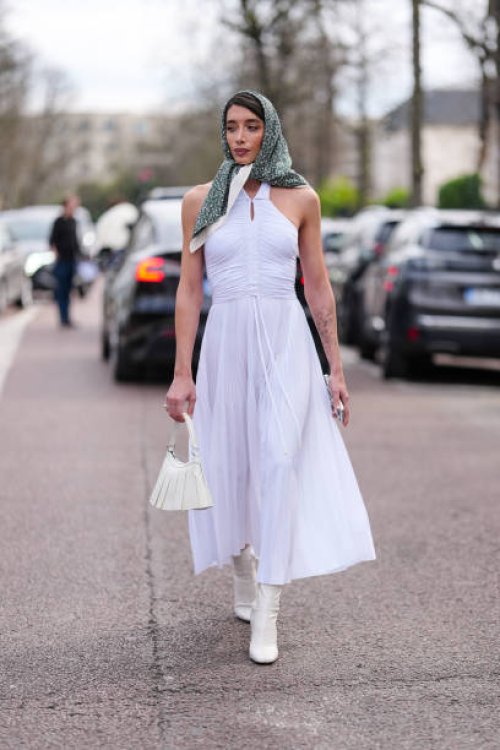 Penelope Guerrero wears black sunglasses, dark green pattern/print head scarf, white halter pleated maxi dress, cream leather bag, white heeled boots...