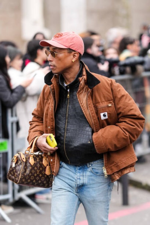 Pharrell Williams wears a light red cap, a brown oversized Sacai jacket, gold necklace, a black leather zipped jacket, a brown leather Louis Vuitton...