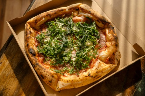pizza with prosciutto and arugula leaves in box on table at home in bright sunlight. take-out comfort food concept. - junk food stock pictures, royalty-free photos & images