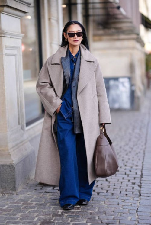 Pornwika Spiecker wears sunglasses , earrings, a gray long wool / trench coat, a black leather jacket , blue flared denim jeans pants, a large...