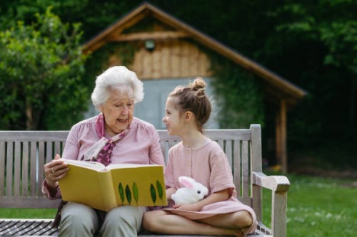 portrait granddaughter with grandmother, sitting on bench in the garden, reading book together. young girl enjoying together time with elderly grandma, looking at photos from photo album. - garden decoration photos et images 