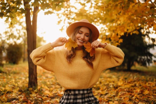 portrait of a beautiful young woman in a hat and a yellow sweater having fun and holding an autumn leaf enjoying solitude walking through the foliage in an autumn park outdoors in autumn. a pretty smiling girl in fashionable 