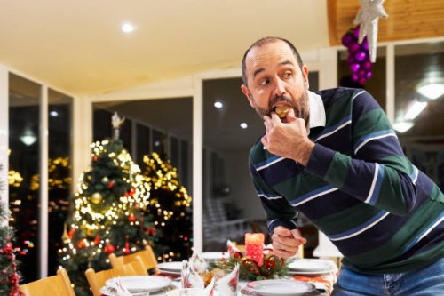 portrait of a man in casual clothes standing making a prank eating a sweet hidden looking away at christmas dinner, front view - food stock pictures, royalty-free photos & images