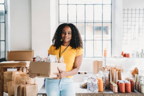 portrait of a mixed race volunteer woman holding a cardboard box of food and drink at the food bank - food stock pictures, royalty-free photos & images