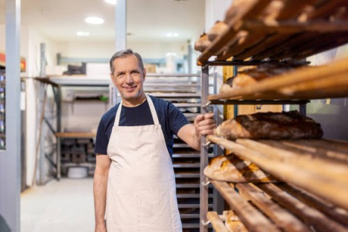 portrait of a senior baker in apron standing by bread rack - food stock pictures, royalty-free photos & images