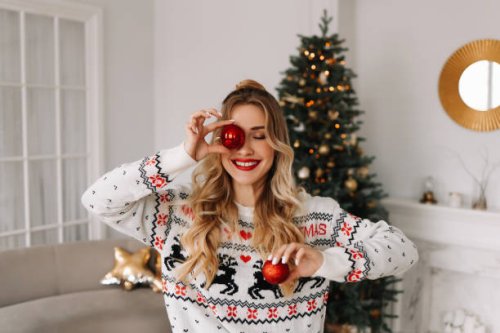 portrait of a young cheerful woman with red lips and curly hair in a knitted sweater laughing and holding a red christmas ball covering her eyes with it against the background of a christmas decorated christmas tree at a holi