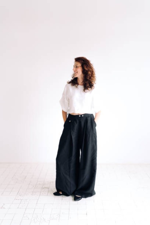 portrait of laughing beautiful authentic young woman with curly brown hair and brown eyes standing against white background with hands in pants pockets. she is wearing loose linen casual clothing. concept of natural beauty - 