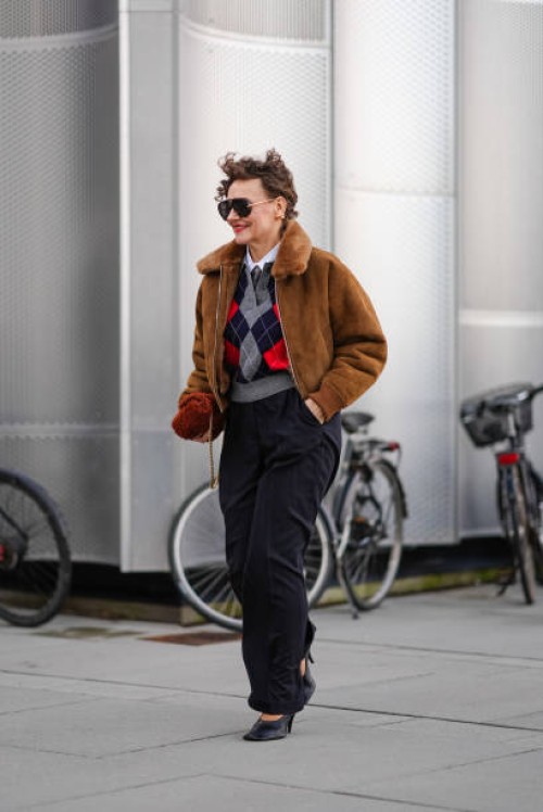 Renia Jaz wears aviator sunglasses, earrings, a brown aviator jacket with fluffy inner lining , an argyle pattern colored pullover , suit flared...