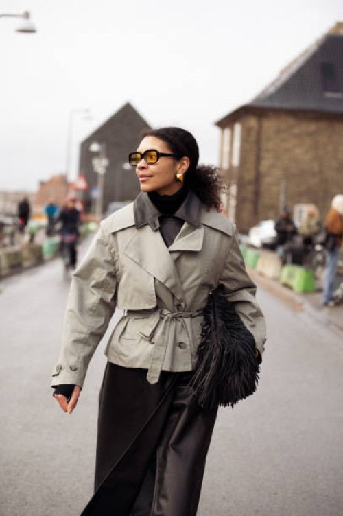 Sara Flaaen Licius wears army green trench crop coat, black leather skirt, orange sunglasses, black leather bag with tassels outside the Munthe...