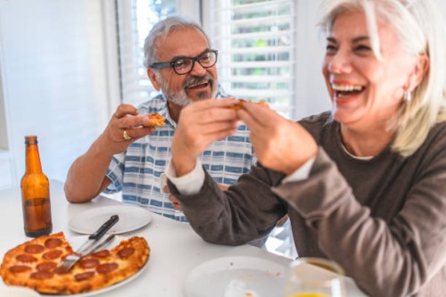senior couple laughing while eating pizza at home - junk food stock pictures, royalty-free photos & images