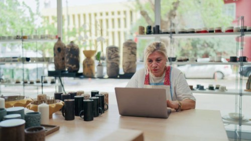 senior female potter working on laptop in her pottery shop - home decoration stock-fotos und bilder