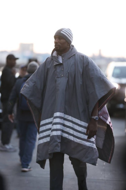Serge Ibaka wears a wide grey blanket jacket with white stripes, matching grey hat, a white t-shirt and grey suit pants, outside before Thom Browne...