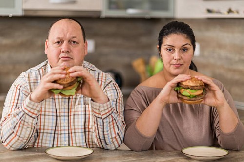 serious overweight couple eating burgers in the kitchen. - junk food stock pictures, royalty-free photos & images