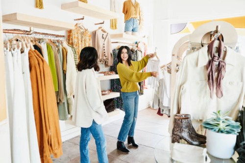 shop owner helping smiling woman choose outfit while shopping in clothing boutique - fashion photos et images de collection