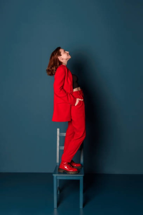 side view of a smiling young woman in a red suit posing standing on a chair in a new clothing collection studio shot indoors - fashion fotografías e imágenes de stock