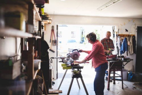 side view of senior woman using circular saw while man holding drill in background - home decoration stock pictures, royalty-free photos & images
