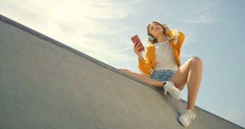 skatepark, headphones and woman on a phone listening to music for calm, gen z summer holiday on blue sky. young person with streetwear and cellphone, smartphone or 5g audio tech in urban city below - fashion stock pictures,