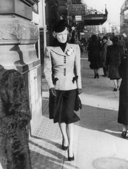 Smartly dressed woman walks down a London street. Her tailored suit has squared shoulders and the contrasting buttons and trim on her jacket match...