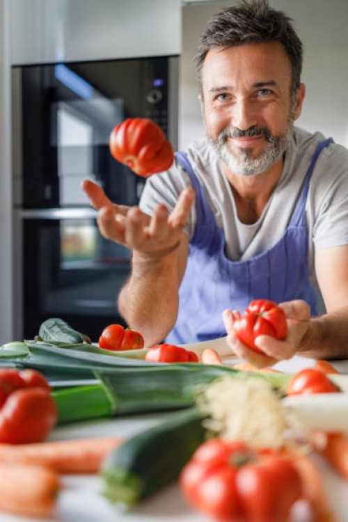 smiling 50 year old man with a beard in front of a table full of fresh vegetables tossing a tomato. healthy food concept. - food stock pictures, royalty-free photos & images