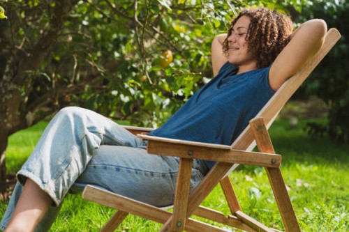 smiling curly haired woman with eyes closed resting on deck chair in garden - garden decoration photos et images de collection
