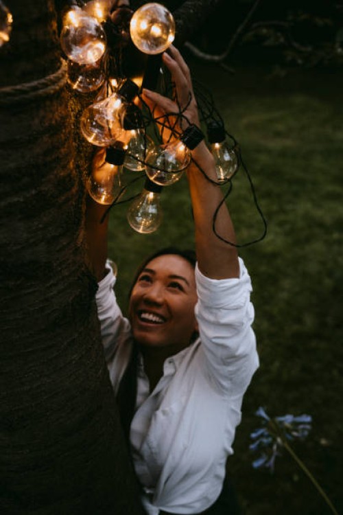 smiling female hanging lighting equipment on tree trunk in yard during dinner party - garden decoration stock pictures, royalty-free photos & images