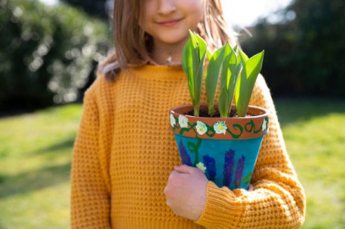 smiling girl (8-9) holding a self-painted terracotta pot filled with young tulip shoots outdoors on a sunny day in springtime. - garden decoration stock pictures, royalty-free photos & images