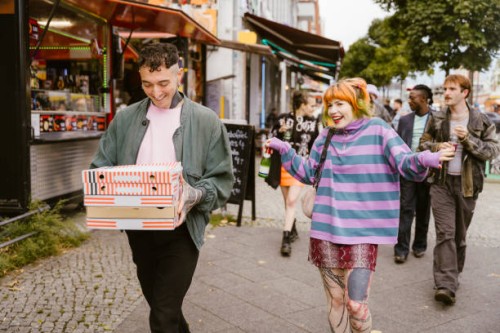 smiling non-binary person holding stack of pizza boxes while walking with friends at street in city - junk food stock pictures, royalty-free photos & images