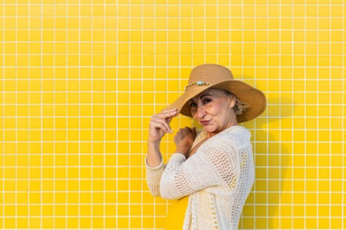smiling senior woman holding her summer hat on a yellow tiled wall - fashion stock pictures, royalty-free photos & images