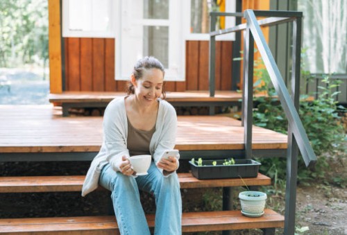 smiling woman with curly hair in casual clothes with cup of morning coffee using mobile phone sitting at backyard of house - garden decoration photos et images de collection
