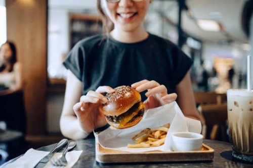 smiling young asian woman enjoying freshly made delicious burger with fries and a glass of iced coffee in a cafe - junk food stock pictures, royalty-free photos & images