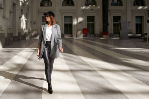 stylish woman wearing white t-shirt, long blazer, black leather pants and black cap walking alone through a public corridor. - fashion photos et images de collection