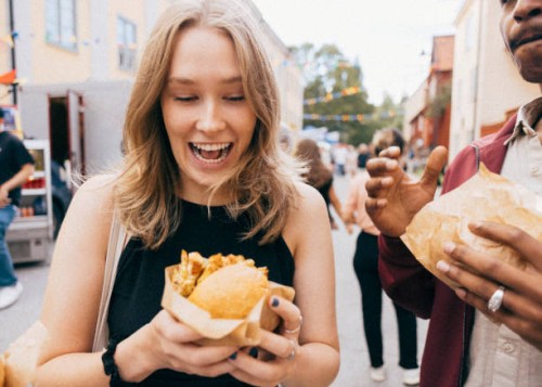 surprised young woman holding burger next to male friend at food festival - junk food stock pictures, royalty-free photos & images