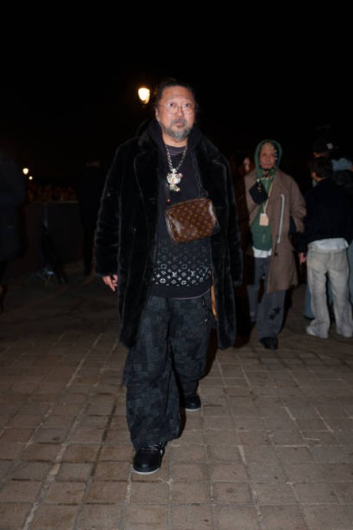 Takashi Murakami wears glasses, a black fluffy coat, a necklace depicting a cartoon character, a black pullover with silver shiny Louis Vuitton...