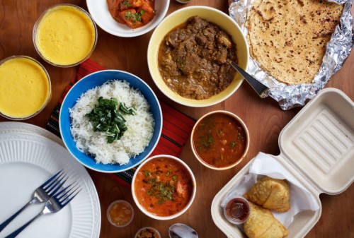 Takeout food from Indigo in Washington, DC: from left: Mango Lassi, Rice, Lamb Curry, Butter Chicken, Chickpeas, Papadam, Samosas, At bottom are...