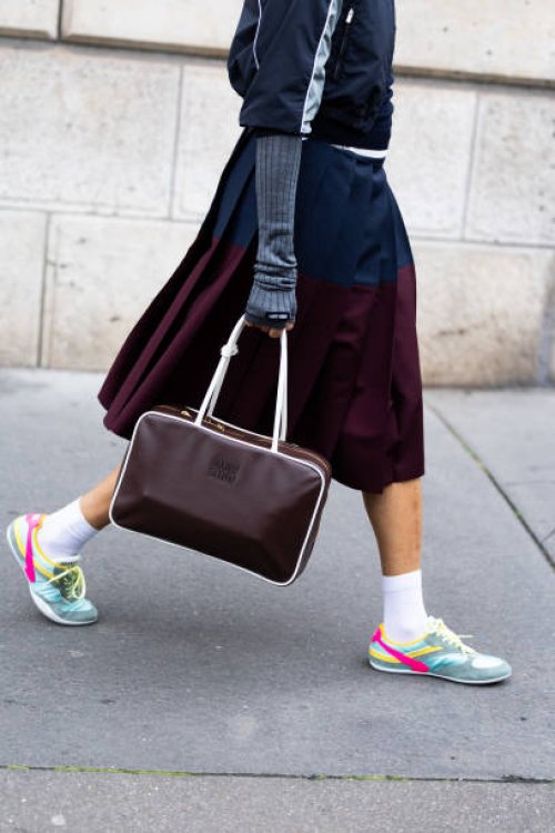 Tamu Mcpherson wears a blue sweater, a grey and dark grey jacket, grey long gloves, a blue and bordeaux pleated maxi skirt, a brown leather Miu Miu...