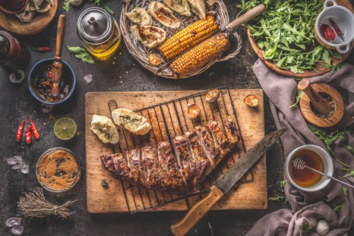 tasty grilled bbq meat ribs on wooden cutting board and grill grate with knife and fresh garlic herb butter bread served on rustic table with grilled corn cobs with bbq sauce , salad leaves and other tasty ingredients - food