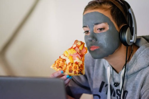 teenage girl wearing headphones and watching her laptop while holding a slice of pizza. - junk food stock pictures, royalty-free photos & images