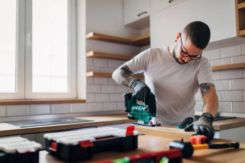 the bearded carpenter using electric jigsaw for cutting a wooden plank in his new kitchen. - home decoration stock pictures, royalty-free photos & images